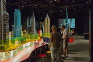 People building with LEGO bricks at a table, surrounded by large display models of skyscrapers in the well-lit Sioux City Art Center's Towers of Tomorrow exhibit space.