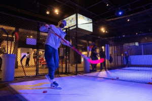 A person practices shooting a hockey puck with a stick inside a netted indoor sports facility in St. Louis, with others participating in the background during an international tour.