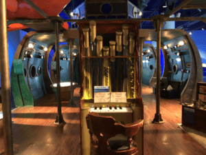 A wooden organ with pipes and a chair stands before the metallic submarine-themed Voyage to the Deep - Underwater Adventures traveling exhibition, complete with round windows and control panels inspired by Jules Verne.