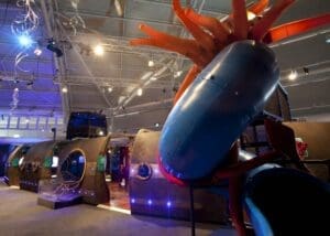 Large indoor playground structure with tubes, slides, and orange tentacle-like features inside a well-lit building, part of the Milwaukee Public Museum's "Voyage to the Deep" underwater exploration exhibit, with interactive displays visible.