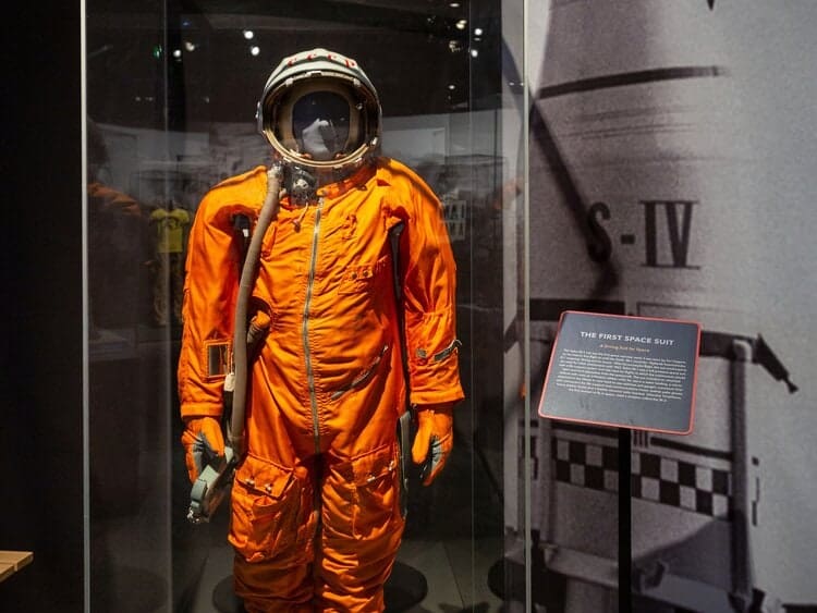 An orange space suit is displayed in a museum case with an informational plaque titled "The First Space Suit," featured as part of the traveling exhibition Apollo: When We Went to the Moon.
