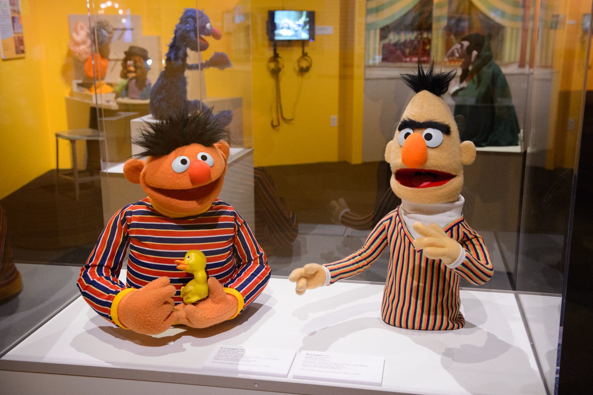 Two puppets resembling Ernie and Bert from Sesame Street are displayed in a glass case at The Jim Henson Exhibition: Imagination Unlimited, with Ernie holding a rubber duck and other museum exhibits visible in the background.