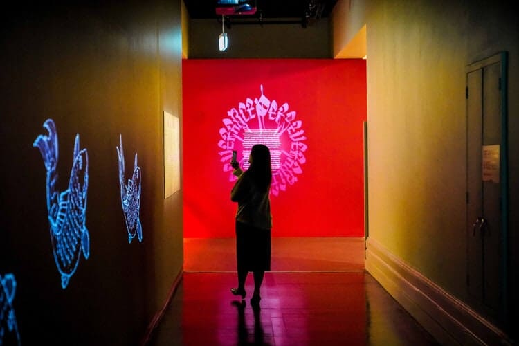A person stands in a dimly lit hallway taking a photo of a bright red and white digital art display projected on the far wall, capturing striking illuminated images along the side walls—perfect for a traveling exhibition photography case study.