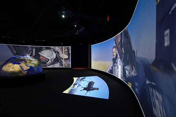 Curved screens display images of astronauts and Earth in a dark exhibit space, with a 3D globe in the center—evoking the wonder of Neil Armstrong on the moon and historic moon landing videos.
