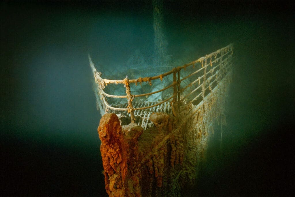 Bow of the sunken Titanic shipwreck underwater, explored by Bob Ballard, covered in rust and marine growth, with railings and metal structure visible in dim light.