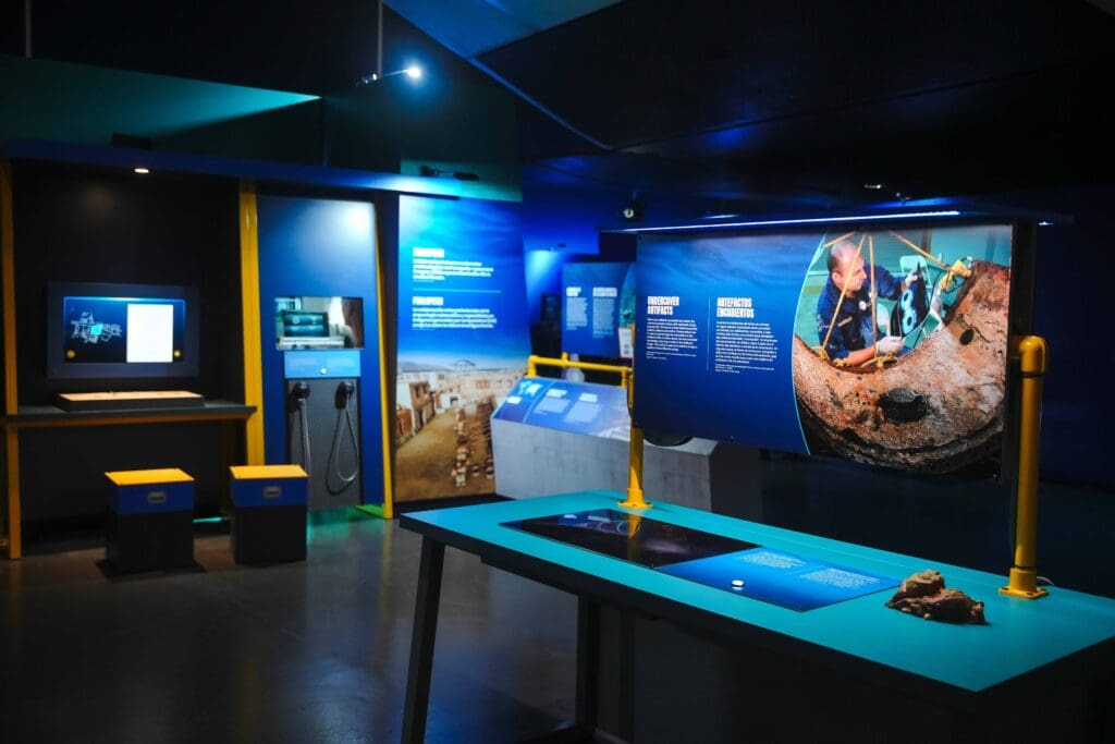A modern museum exhibition, featured by the Smithsonian, features informational panels, photographs, interactive displays, and artifacts from Mysteries from the Deep under bright blue lighting.