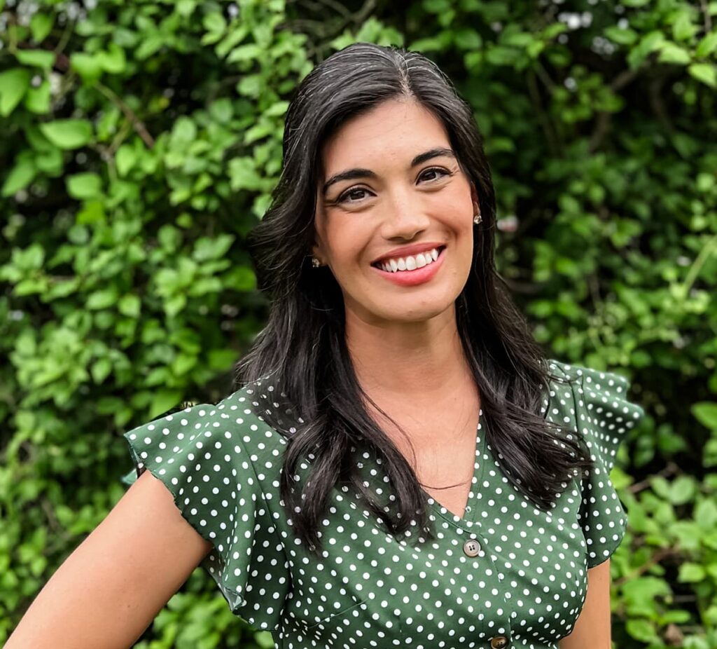 Woman with long dark hair wearing a green polka dot dress smiles while standing outdoors in front of leafy greenery.
