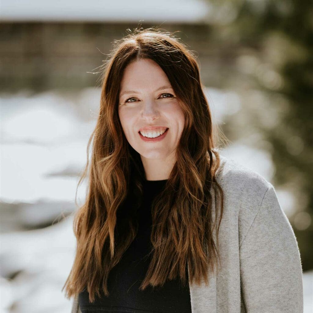 A woman with long brown hair, wearing a grey cardigan and black top, smiles outdoors with a blurred background.