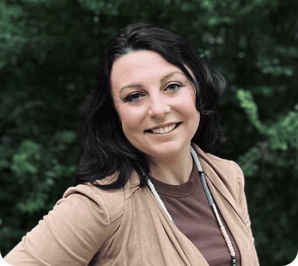 Jeanette Collins, a woman with long dark hair and a light brown sleeveless cardigan, smiles outdoors with lush greenery in the background.