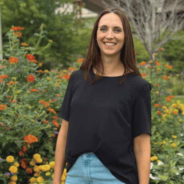 Liz Frerichs, a woman with long brown hair wearing a black shirt and light blue jeans, stands smiling in front of a garden filled with various colorful flowers.