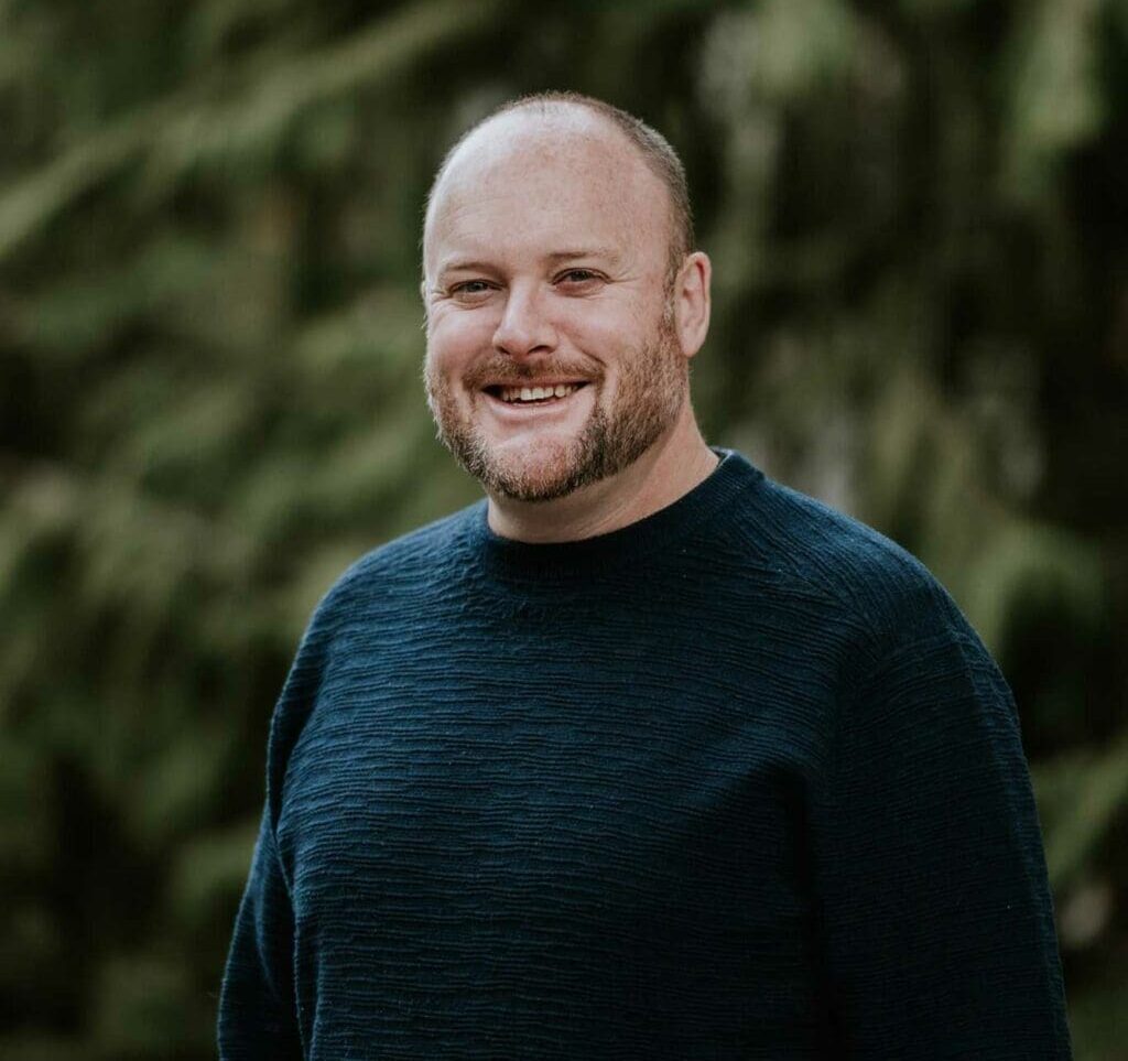 A man with a beard and short hair, wearing a dark blue sweater, stands outdoors in front of blurred greenery, smiling at the camera.