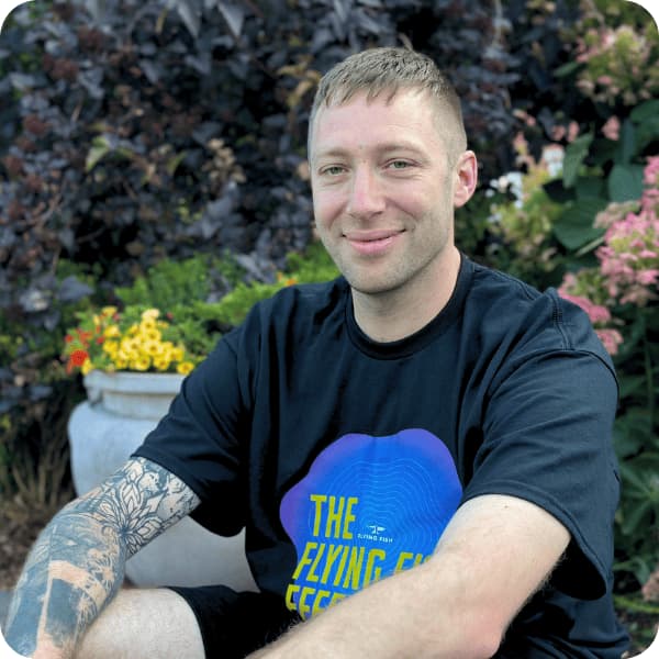Ziggy Goddard, a man with short hair and a tattooed arm, sits outdoors smiling in a black T-shirt with colorful text. Flowers and green plants create a vibrant backdrop.