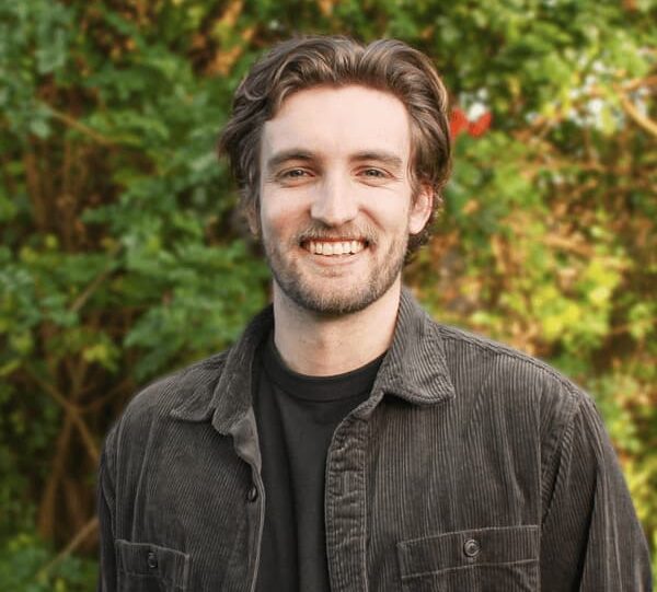 Kai Lane-U'ren, a man with wavy brown hair and a beard, smiles outdoors, wearing a dark corduroy shirt over a black t-shirt, with green foliage in the background.