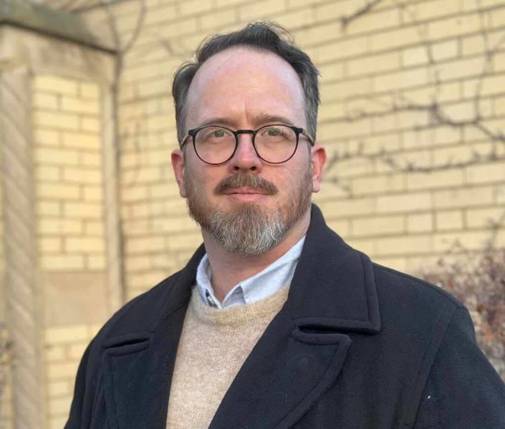 Joseph Mandrell, a man with glasses and a beard, stands outdoors in front of a yellow brick wall, wearing a black coat over a tan sweater and collared shirt.