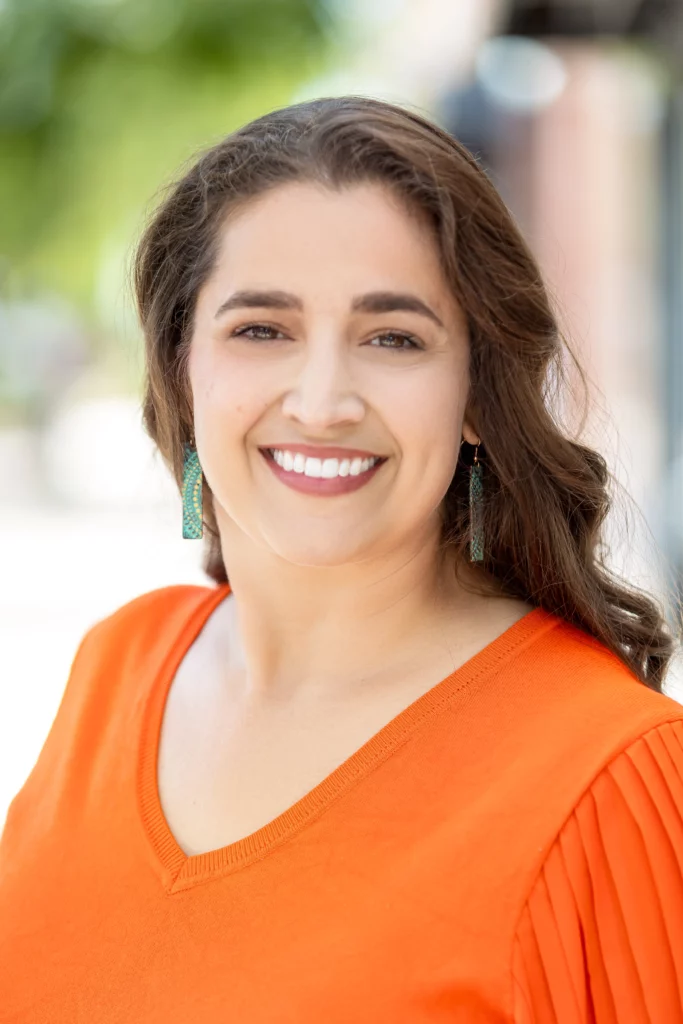 Emily Kielhorn, a woman with long brown hair wearing an orange top and teal earrings, smiles outdoors with a blurred background.