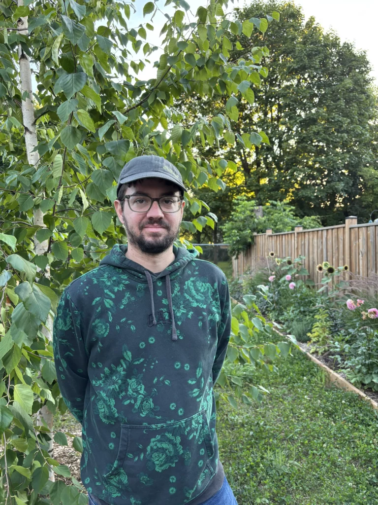 Daniel MacPherson, wearing glasses, a dark cap, and a green patterned hoodie, stands in a garden surrounded by trees, flowers, and a wooden fence in the background.